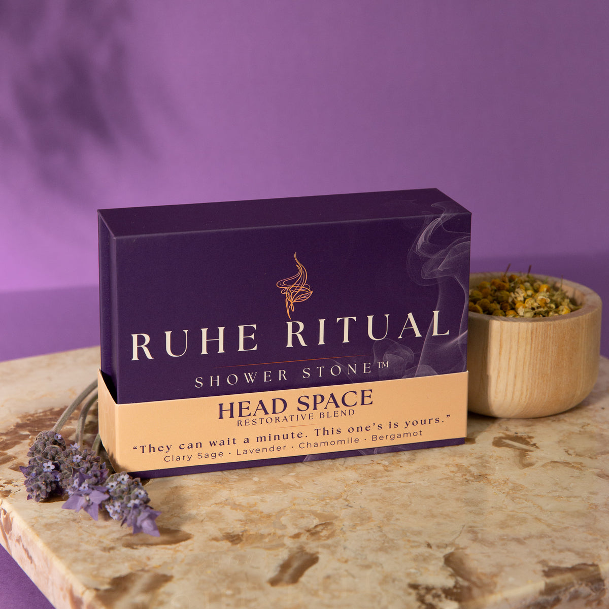 A box of Ruhe Ritual Shower Stone Head Space restorative blend placed on a stone surface, with dried lavender sprigs and a small wooden bowl of loose botanicals nearby against a purple background.
