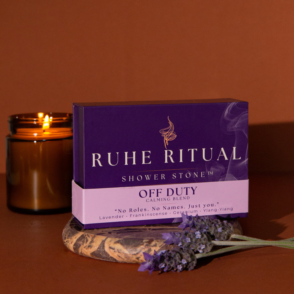 Ruhe Ritual Shower Stone 'Off Duty' box in a purple and lavender color scheme, styled on a wooden coaster next to a lit amber candle and lavender sprigs against a warm brown background.