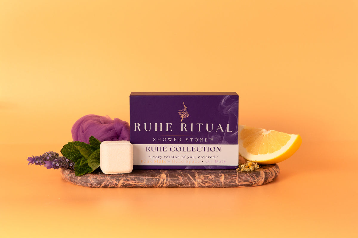 A boxed shower stone product from Ruhe Ritual sits on a stone tray with a white cube-shaped stone, purple moss, green leaves, lavender sprigs, and a lemon slice against a warm orange background.